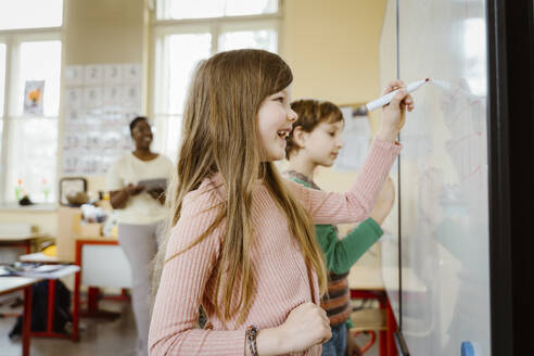 Happy girl writing on whiteboard while solving maths problem in classroom - MASF37275