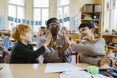 Happy teacher and boys giving high-fives while sitting at desk in classroom - MASF37294