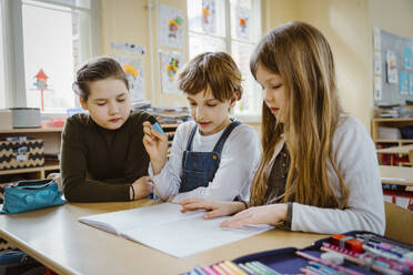 Boy and girls sharing book while studying together at desk in classroom - MASF37296