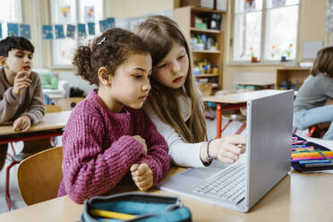 Schoolgirls sharing laptop together at desk in classroom - MASF37300