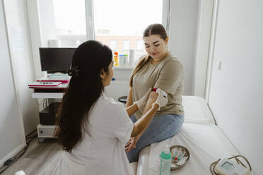 Doctor tying band on patient's arm sitting on bed in clinic - MASF37308