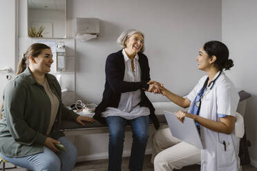 Happy senior woman shaking hand with doctor sitting in clinic - MASF37321