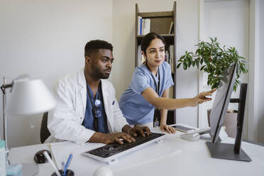 Doctor discussing with male coworker working on computer at desk in clinic - MASF37333