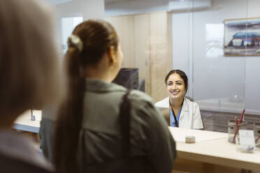 Smiling receptionist looking at woman through glass shield in hospital - MASF37345