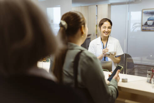 Happy female receptionist looking at woman through glass window at hospital - MASF37347