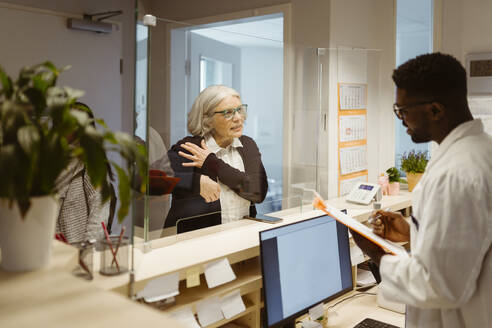 Senior female patient explaining health issues to male receptionist through transparent shield in clinic - MASF37352