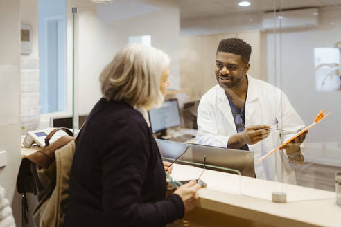 Smiling male receptionist holding clipboard while talking to senior woman through transparent shield in clinic - MASF37354