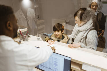 High angle view of smiling boy passing document to male receptionist below transparent shield in clinic - MASF37358