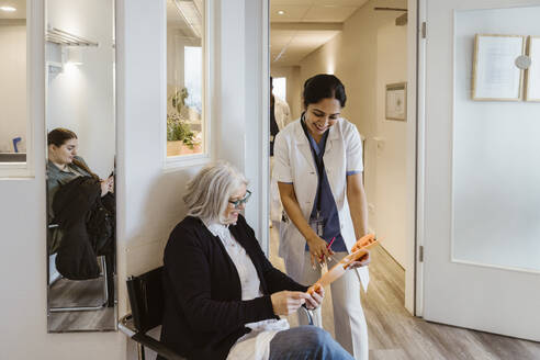 Smiling female healthcare worker standing by senior patient looking at clipboard in clinic - MASF37371