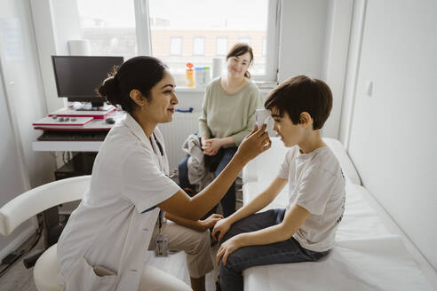 Smiling female pediatrician measuring body temperature of boy through infrared thermometer at clinic - MASF37382