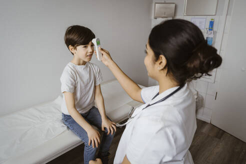Female pediatrician measuring temperature of boy through infrared thermometer at hospital - MASF37383