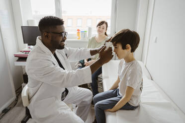 Male pediatrician examining boy sitting on bed in examination room at healthcare center - MASF37384