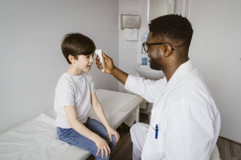 Young male pediatrician measuring temperature of boy using infrared thermometer at clinic - MASF37385