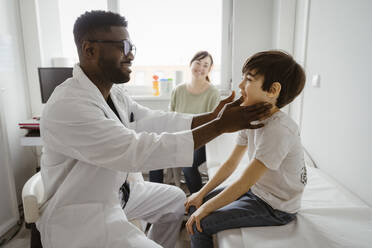 Smiling male pediatrician examining neck of boy sitting on bed at healthcare center - MASF37386