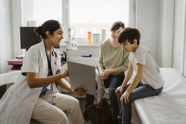 Female pediatrician showing tablet PC to mother and son sitting in examination room at clinic - MASF37389