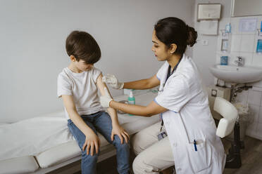 Female pediatrician disinfecting boy's arm sitting on bed at healthcare center - MASF37391