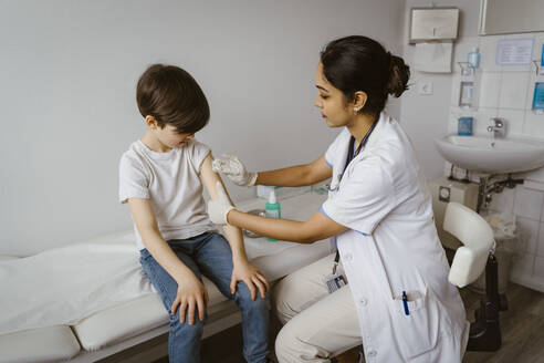 Female pediatrician disinfecting boy's arm sitting on bed at healthcare center - MASF37391