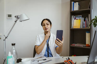 Female doctor making online video call with patient through smart phone while sitting at desk in clinic - MASF37399