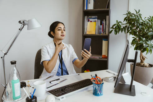 Female physician explaining patient over online video call through mobile phone while sitting at desk in clinic - MASF37400