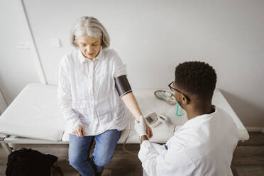 High angle view of male doctor measuring blood pressure of senior patient sitting on bed at medical clinic - MASF37407