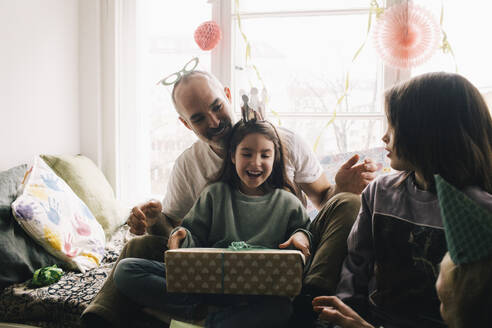 Surprised girl holding birthday present while sitting with brother and father at home - MASF37414