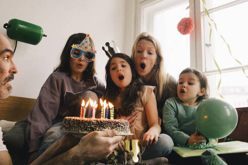 Man holding birthday cake with burning candles in front of surprised family at home - MASF37419