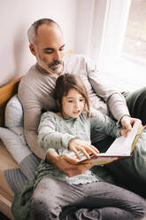 Father and daughter reading book while sitting together on alcove window seat at home - MASF37444