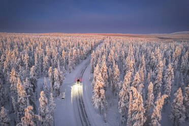 Aerial view of car on icy road and illuminated headlamps driving in the snowcapped forest, at dawn, Akaslompolo, Kolari, Pallas-Yllastunturi National Park, Lapland region, Finland, Europe - RHPLF24503