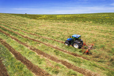 Blue tractor with hay tedder at work on agricultural mowed field, aerial side view, Italy, Europe - RHPLF25546