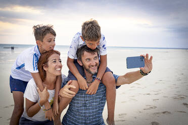 A joyful family of four captures a moment on a Zanzibar beach, smiling for a selfie with their smartphone - RHPLF25627