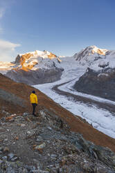 Hiker stands on rocks admiring Monte Rosa massif, Gorner Glacier (Gornergletscher) and Lyskamm peaks at sunset, Riffealp, Zermatt, canton of Valais, Swiss Alps, Switzerland, Europe - RHPLF25731