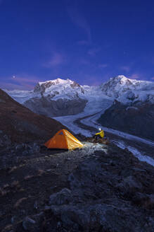 Hiker with head lamp sitting beside the tent in front of Monte Rosa massif, Gorner glacier (Gornergletscher) and Lyskamm peaks at dusk, Riffelsee, Zermatt, canton of Valais, Swiss Alps, Switzerland, Europe - RHPLF25732