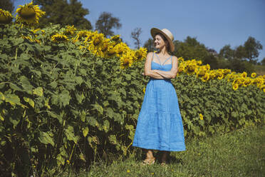 Lächelnde Frau in blauem Kleid in einem Sonnenblumenfeld stehend - NDEF00670