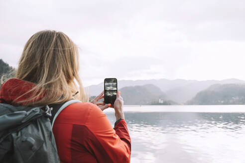 Frau mit Rucksack, die in der Nähe eines Sees ein Foto mit ihrem Smartphone macht - NDEF00684