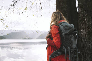 Frau mit Rucksack steht neben einem Baum in der Nähe eines Sees - NDEF00686