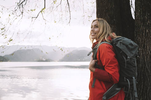 Lächelnde Frau mit Rucksack steht neben einem Baum in der Nähe eines Sees - NDEF00687