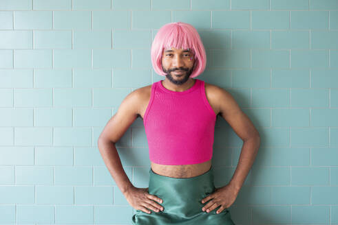 Young queer man standing with arms akimbo in front of tiled wall - VRAF00122