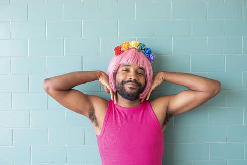 Smiling queer man with flowers on head in front of tiled wall - VRAF00124