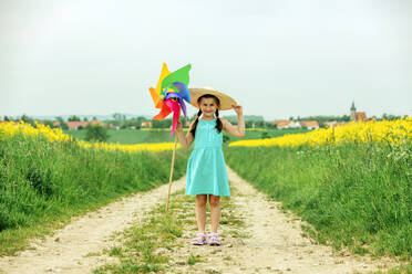 Happy girl wearing hat standing with pinwheel toy standing at field - VSNF00958