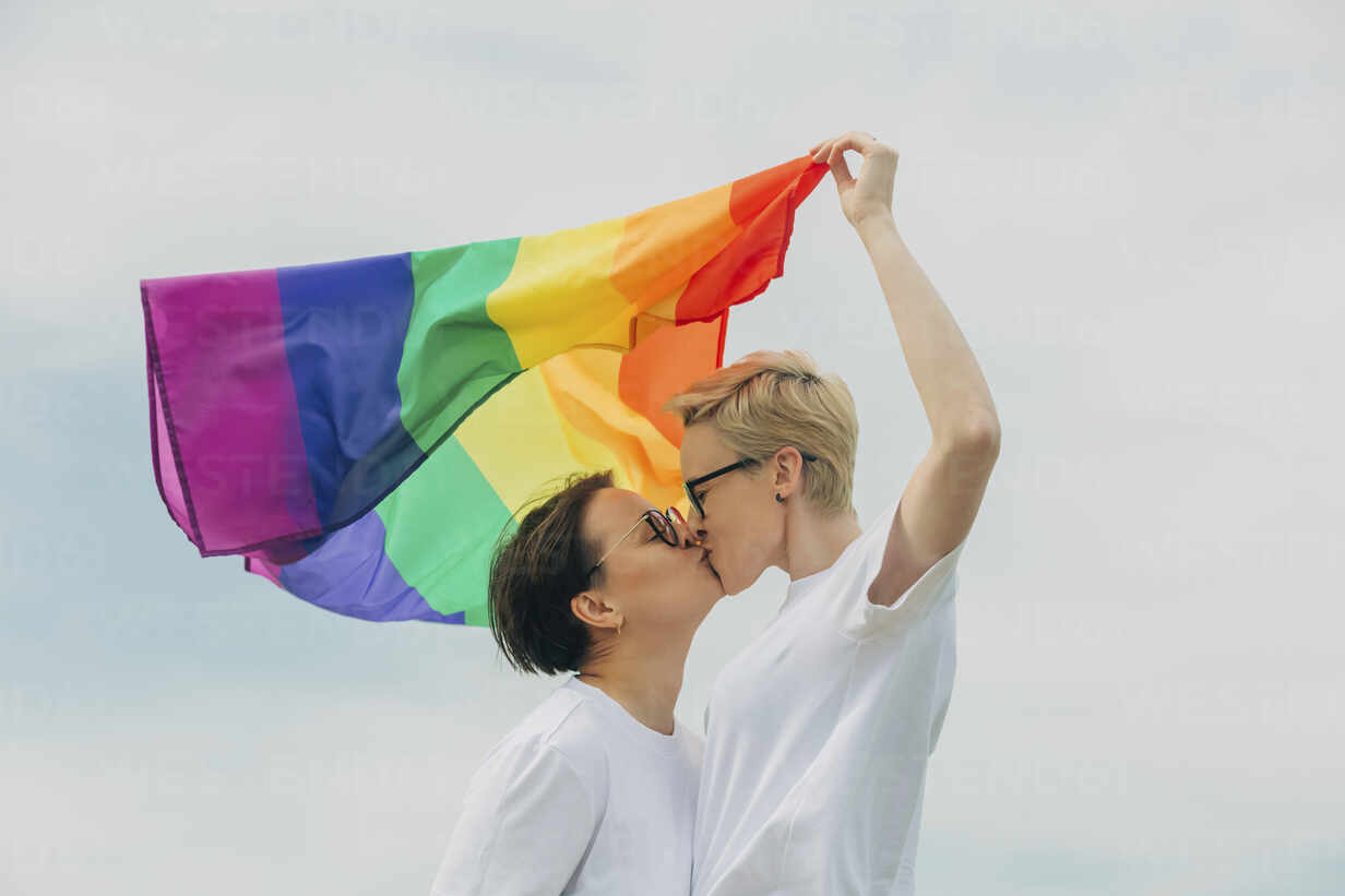 Loving lesbians with rainbow flag kissing each other under sky stock photo