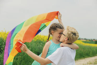 Happy lesbian mother holding rainbow flag and kissing daughter at field - VSNF00979