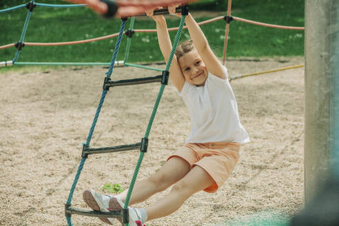 Lächelndes Mädchen klettert auf dem Spielplatz auf eine Strickleiter - VSNF01010