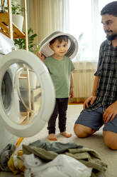 Smiling boy helping father washing clothes in machine at home - ANAF01496