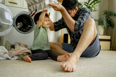 Happy father and son having fun with laundry basket and washing clothes in machine at home - ANAF01497