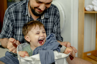 Cheerful boy sitting in laundry bag and having fun with father at home - ANAF01513