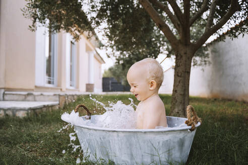 Baby boy taking bath and splashing water in tub at backyard - NDEF00719