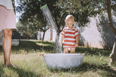 Toddler standing with father pouring water in bathtub at backyard - NDEF00721