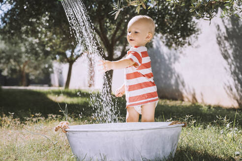 Baby-Junge spielt mit fließendem Wasser in der Nähe der Badewanne stehend - NDEF00722