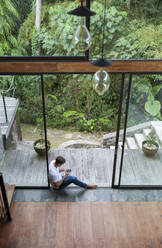 Young man using smart phone sitting by glass door - IKF00733