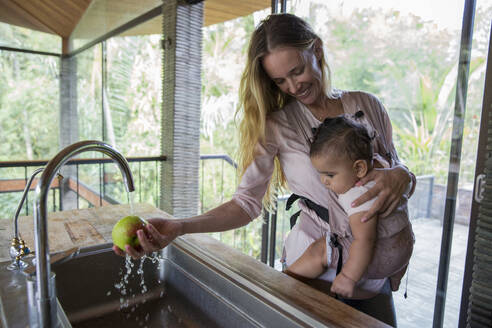 Glückliche Frau wäscht Apfel und trägt Tochter zu Hause - IKF00734